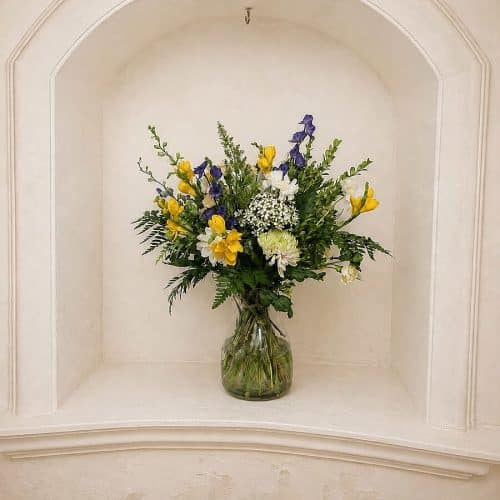 Arreglo floral silvestre Calypso con flores de colores y abundante follaje verde en un jarrón de cristal.